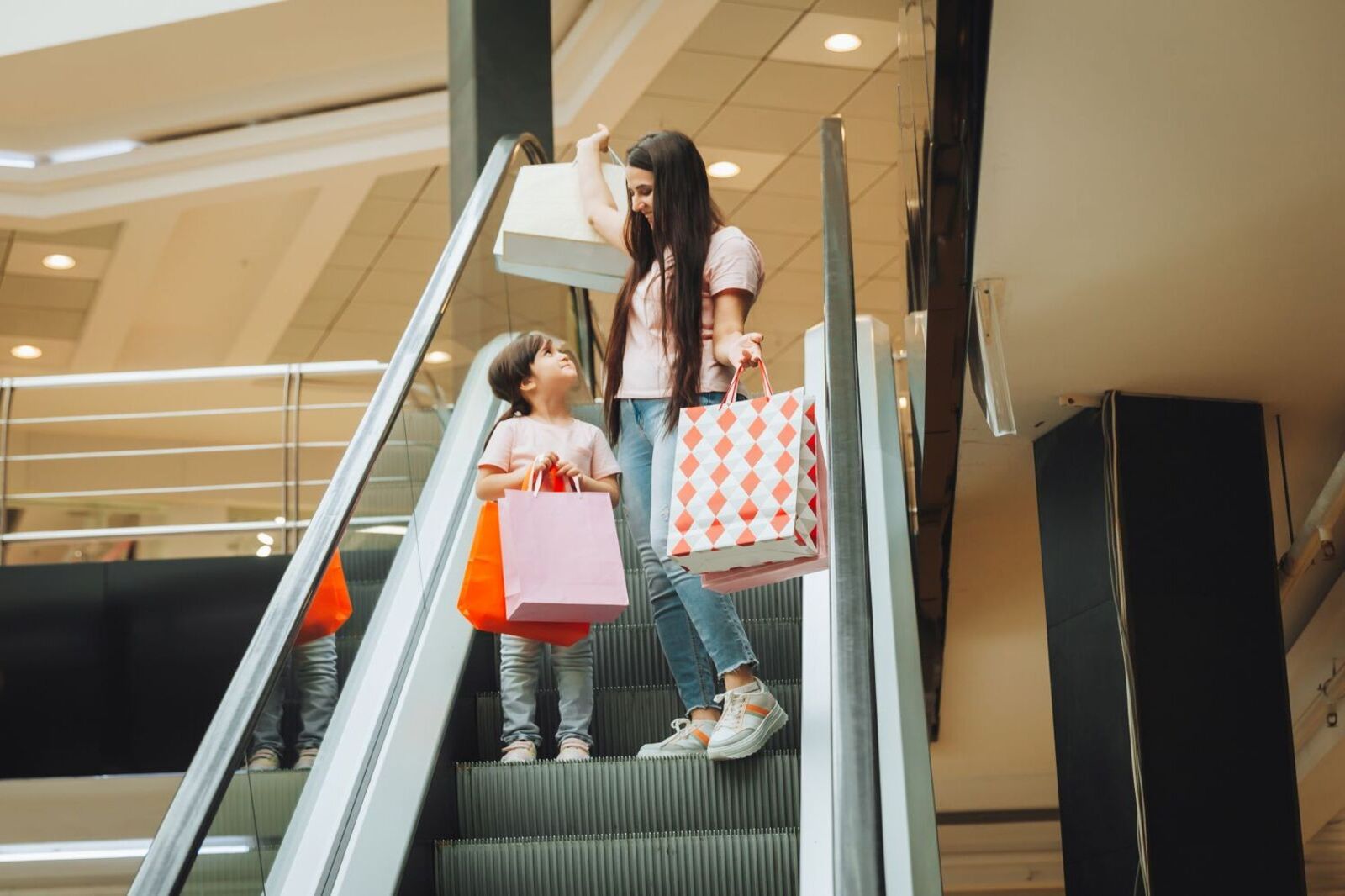 Mãe com criança fazendo compras de dia das mães em shopping com sacolas de presente em uma escada rolante